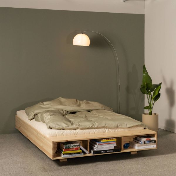 Karup futonbed Merge van FSC-gecertificeerd dennenhout met lage, ‘zwevende’ basis; drie open vakken vol boeken aan het voeteneind en lichtolijfgroen beddengoed in een minimalistische slaapkamer.